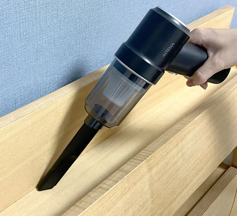 黒いコンパクトなハンディクリーナーを使って、木製の家具の隙間を掃除している様子です。手軽に使えるコードレスタイプで、日常の掃除風景を示しています。