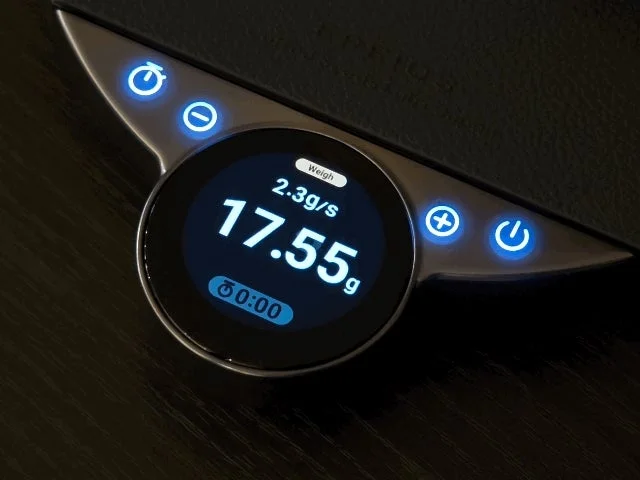 暗い背景に置かれたデジタルスケールのディスプレイが中心に写っている。画面には「Weigh」「17.55g」「2.3g/s」「0:00」と表示され、青く光る操作ボタンが周囲にある。精密な計量やタイマー機能を持つ電子機器の様子を捉えている。
