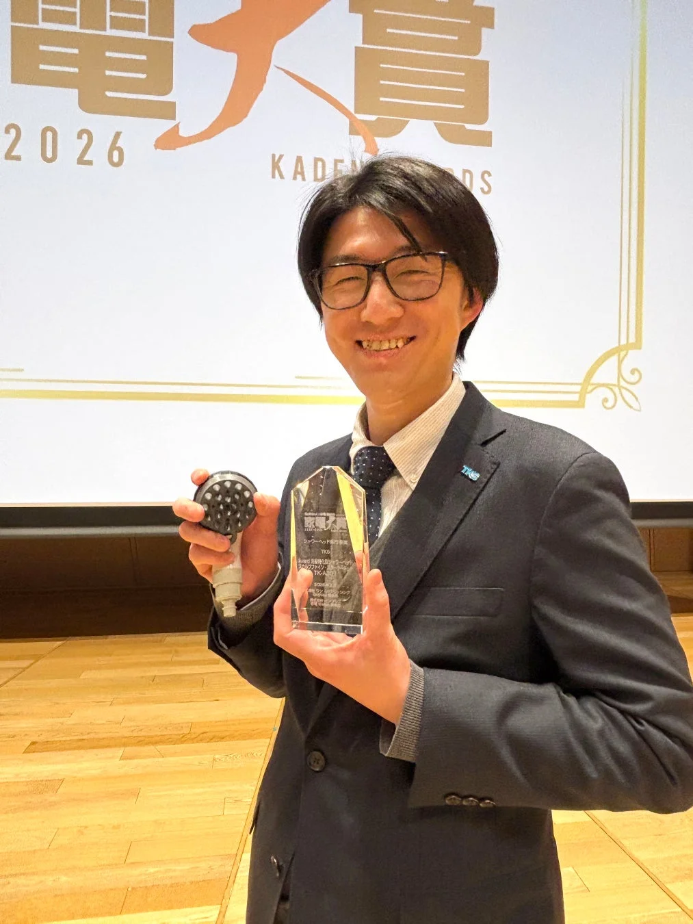スーツ姿の男性が「家電大賞」のトロフィーとシャワーヘッドを手に満面の笑みを浮かべている