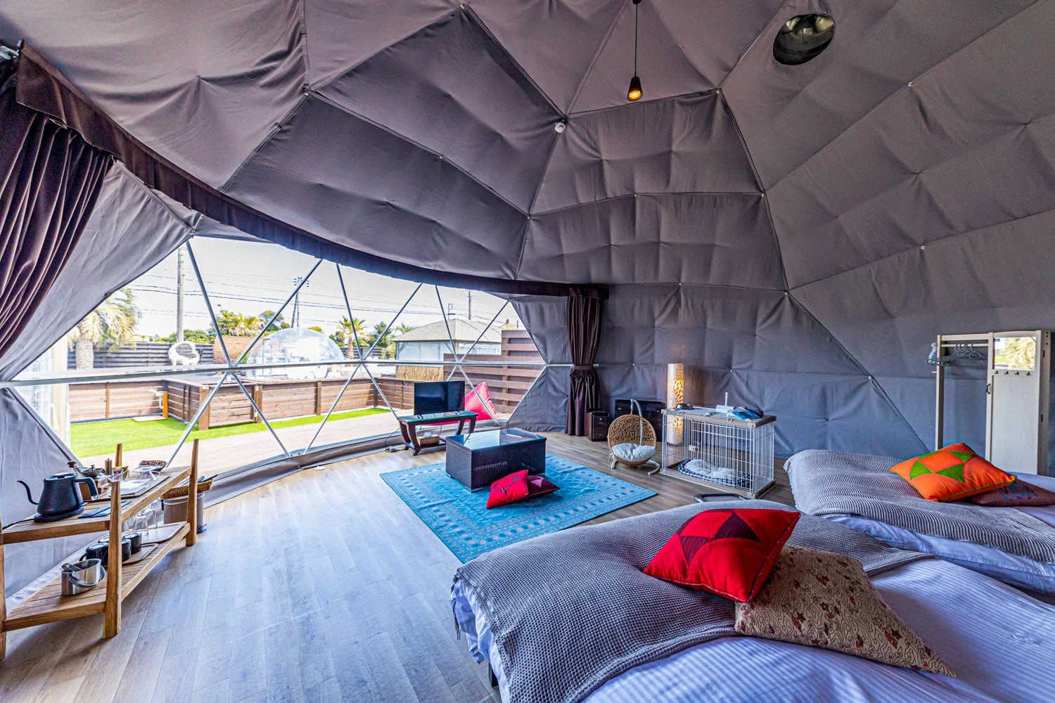 ドーム型テントのモダンな宿泊施設の室内。広々とした空間にはベッド、リビングエリア、ペット用品が配置され、窓からは屋外の庭が見える。快適でペット同伴可能なグランピング施設です。