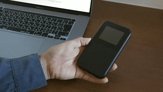 木製のデスクで、ノートパソコンの横に置かれた黒いモバイルデバイス（モバイルルーターまたはスマートフォン）を片手で持っている人物の様子を捉えた画像です。