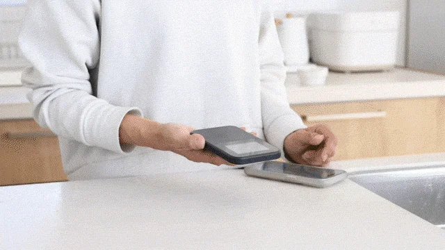 白いカウンタートップの上で、人物が黒いスマートフォンを銀色のスマートフォンの上に置こうとしている様子。キッチンを背景に、デバイス間の操作が行われている場面を示唆しています。