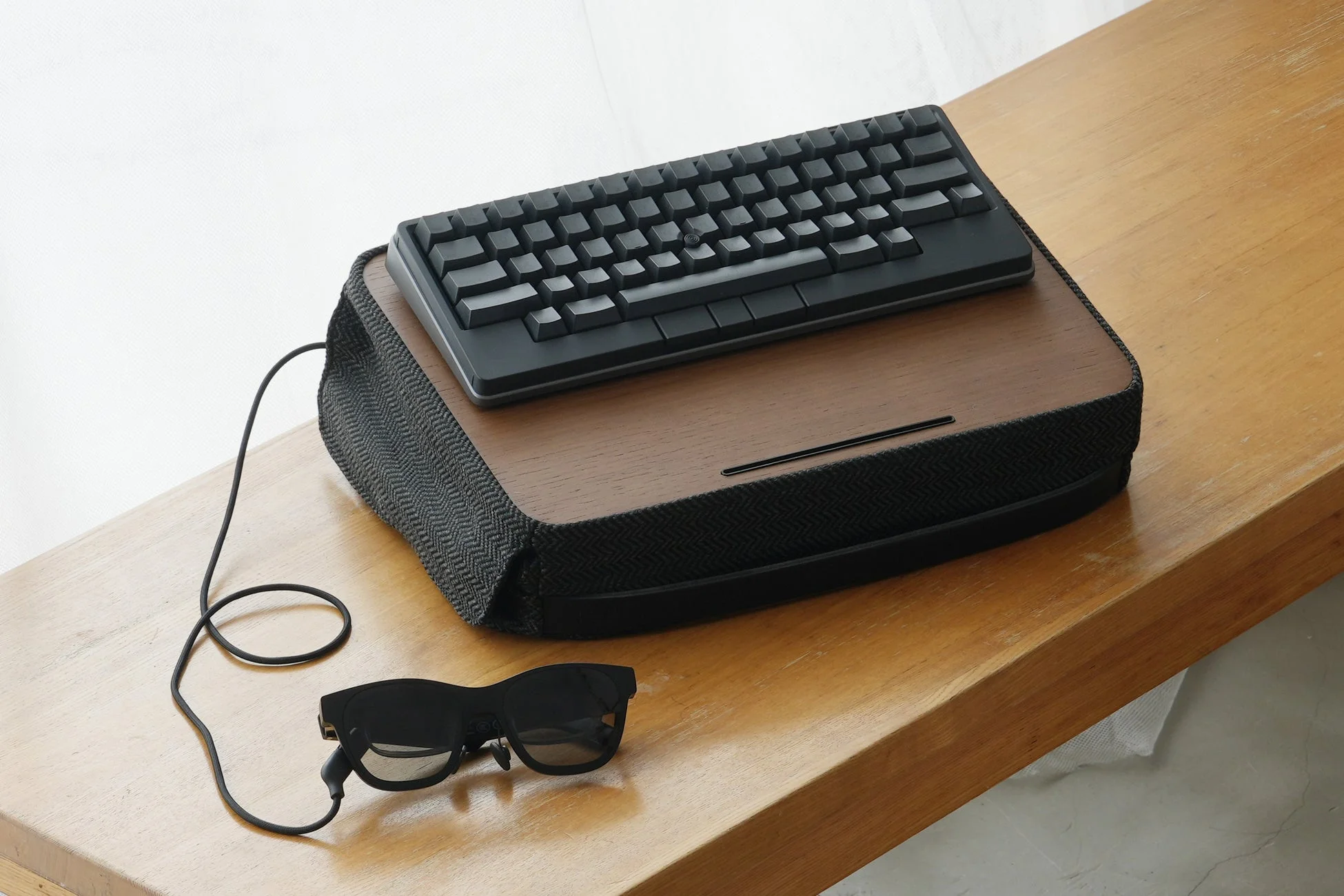 木製の台の上に置かれたミニマルな黒いキーボードと、その横にケーブルで繋がれたスマートグラスのセットアップ。モダンで機能的な電子機器の組み合わせが、すっきりとした空間に配置されています。