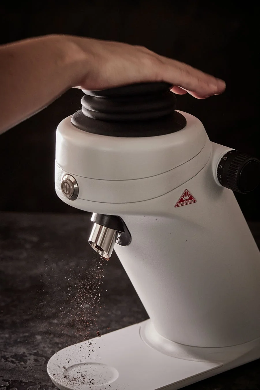 白いマールクーニックのコーヒーグラインダーから挽きたてのコーヒー粉が排出されている様子。手がグラインダーのトップを押している。