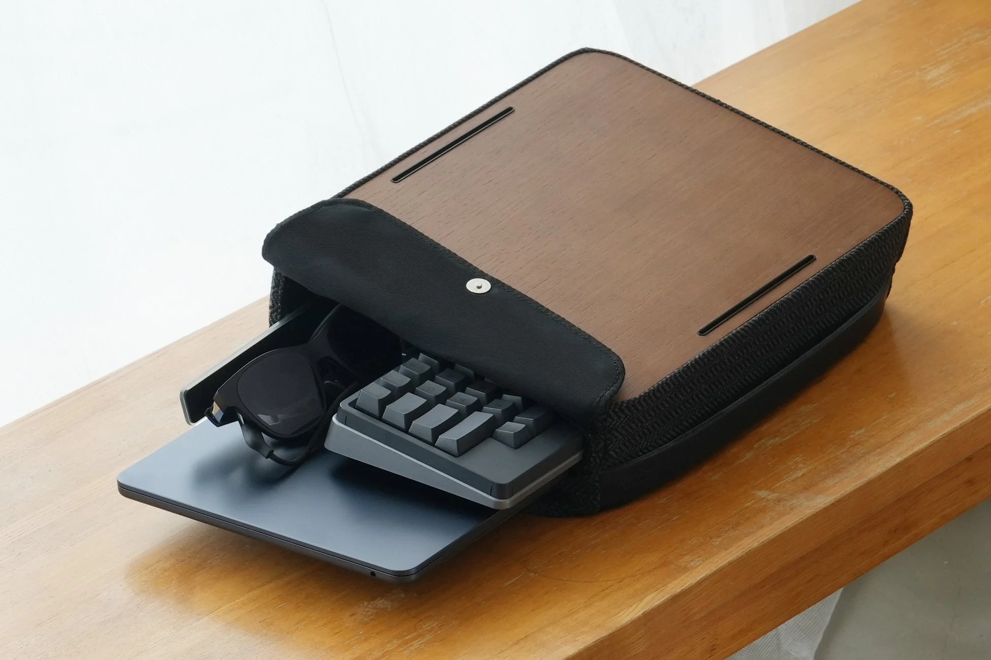 木製の天板を持つスタイリッシュなキャリングケースが木製のテーブルに置かれ、中からノートパソコン、コンパクトなキーボード、サングラスが見えている。現代的なモバイルワーク環境を思わせる収納アイテムの様子。