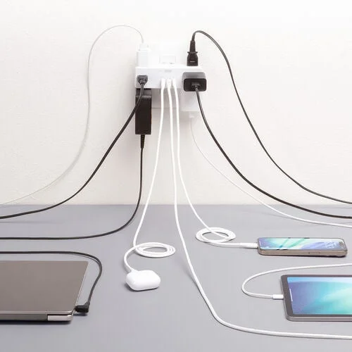 壁の電源タップに複数の充電器が接続され、ノートパソコン、スマートフォン、タブレット、ワイヤレスイヤホンなどの電子機器が同時に充電されている様子。多くのケーブルが絡み合い、現代のデジタルライフにおける充電環境を示しています。