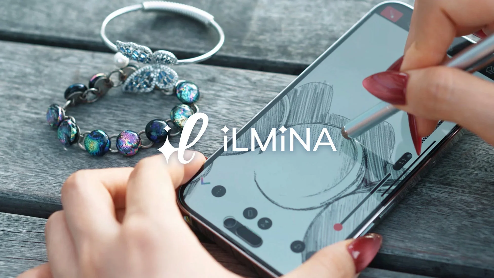 スマートフォンにスタイラスペンで描画している手元と、隣に置かれたきらびやかなブレスレットが写っている。