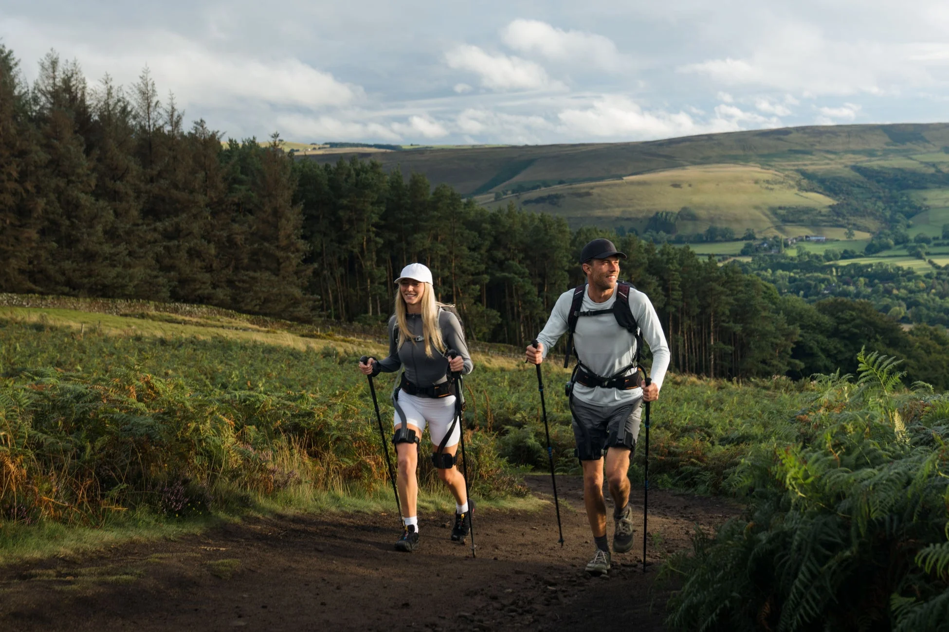 丘陵地帯の未舗装の道を、男女二人がトレッキングポールを使ってハイキングしている様子