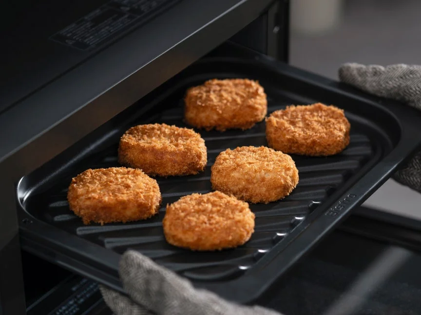 オーブンから、こんがりと揚がったコロッケのような揚げ物が6つ並んだ黒いトレーを取り出している様子