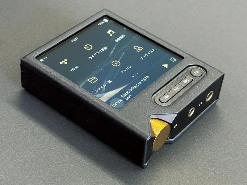 黒い筐体のデジタルオーディオプレーヤー（DAP）が斜め上から撮影されています。画面には音楽再生アプリのインターフェースが表示されており、右側面には物理ボタンと金色のボリュームノブ、オーディオジャックが見えます。
