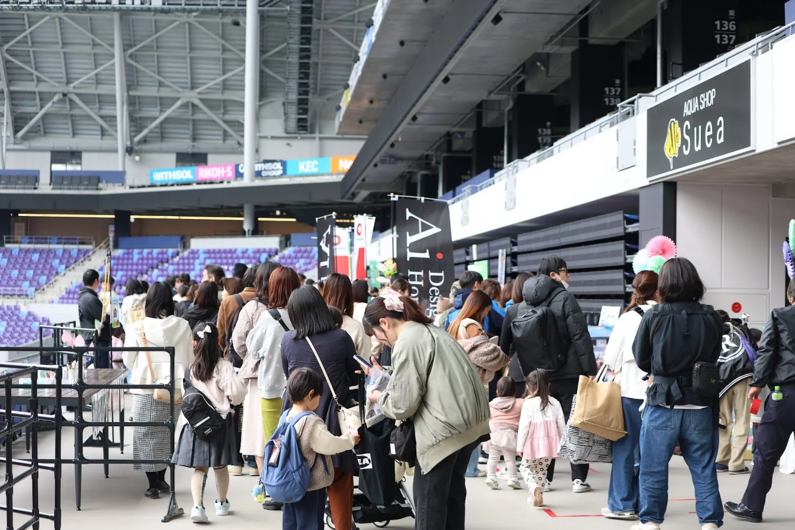スタジアムのコンコースで賑わう屋内イベントの様子。多くの人々、特に家族連れが様々なブースに立ち寄ったり、行列を作ったりしており、活気のある交流やショッピングを楽しんでいる。