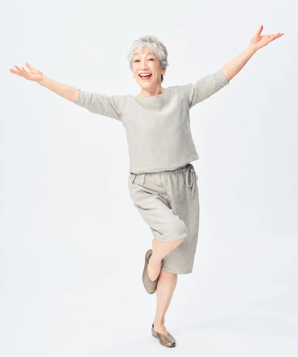 白い背景の前で、短い白髪の高齢女性が、薄いグレーのカジュアルな服装で両腕を広げ、片足を上げて楽しそうに微笑んでいます。活動的で喜びにあふれた表情です。