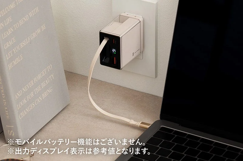 壁のコンセントに接続されたコンパクトな充電器がノートパソコンを充電している様子です。充電器には65Wの出力が表示されており、隣には本が置かれています。