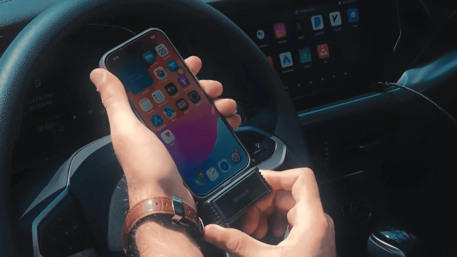 車内でハンドル近くにiPhoneとBelkin製のデバイスを持つ手のクローズアップ。iPhoneの画面には多数のアプリが表示され、車のダッシュボードのディスプレイにもアプリのアイコンが見える。車内でのデジタルデバイスの使用風景です。