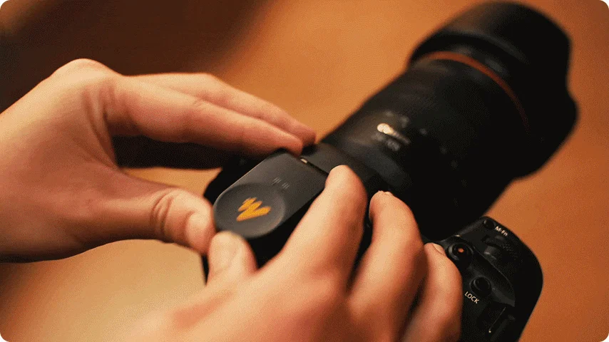 人物が大型レンズを装着した黒いカメラに、黄色のロゴが入った黒いアクセサリーを取り付けている様子