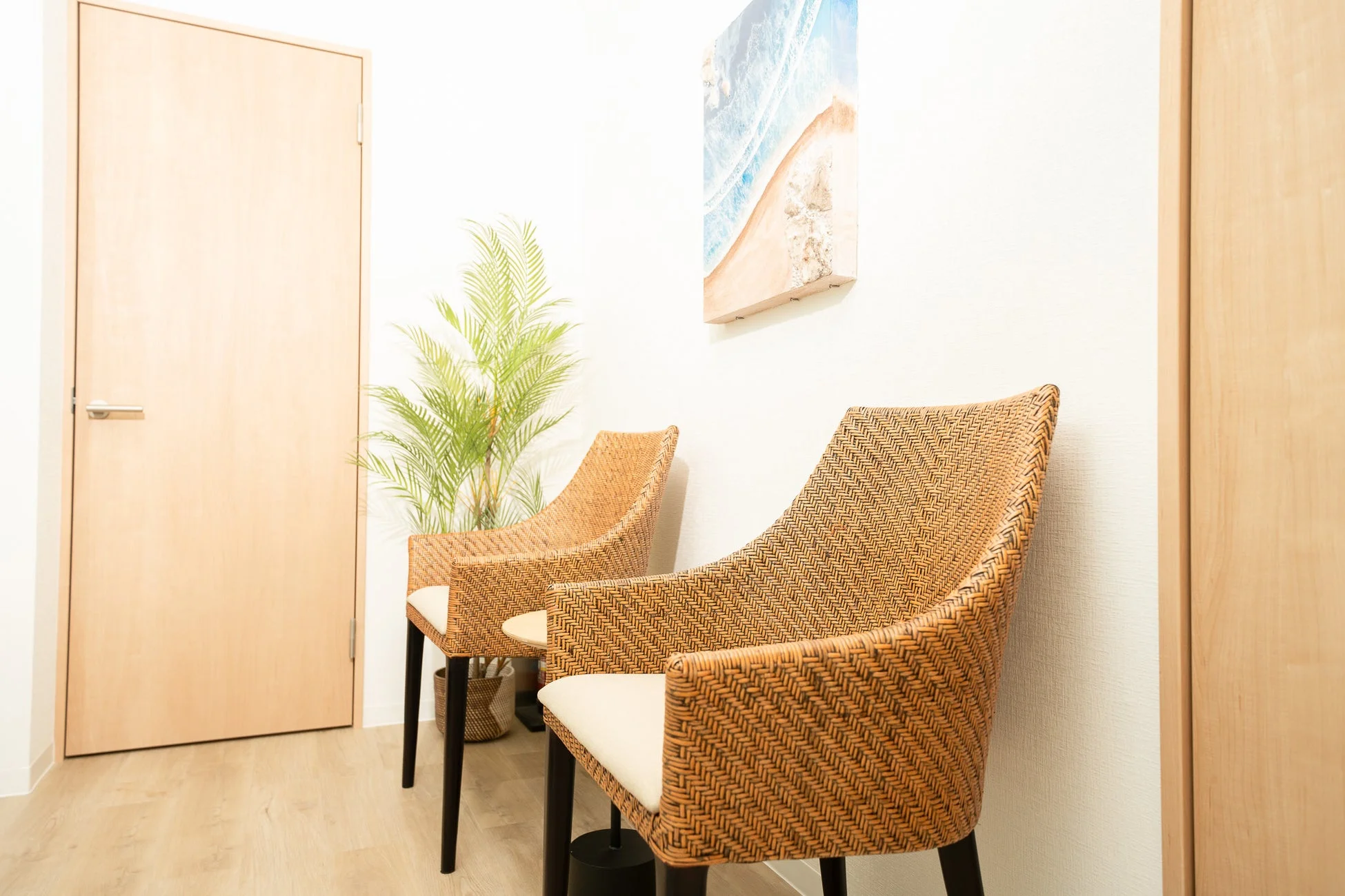 明るい白い壁と木目調の床の空間に、ラタン素材の椅子が2脚並べられています。観葉植物とビーチのアートが飾られ、リラックスできる待合室のような雰囲気です。