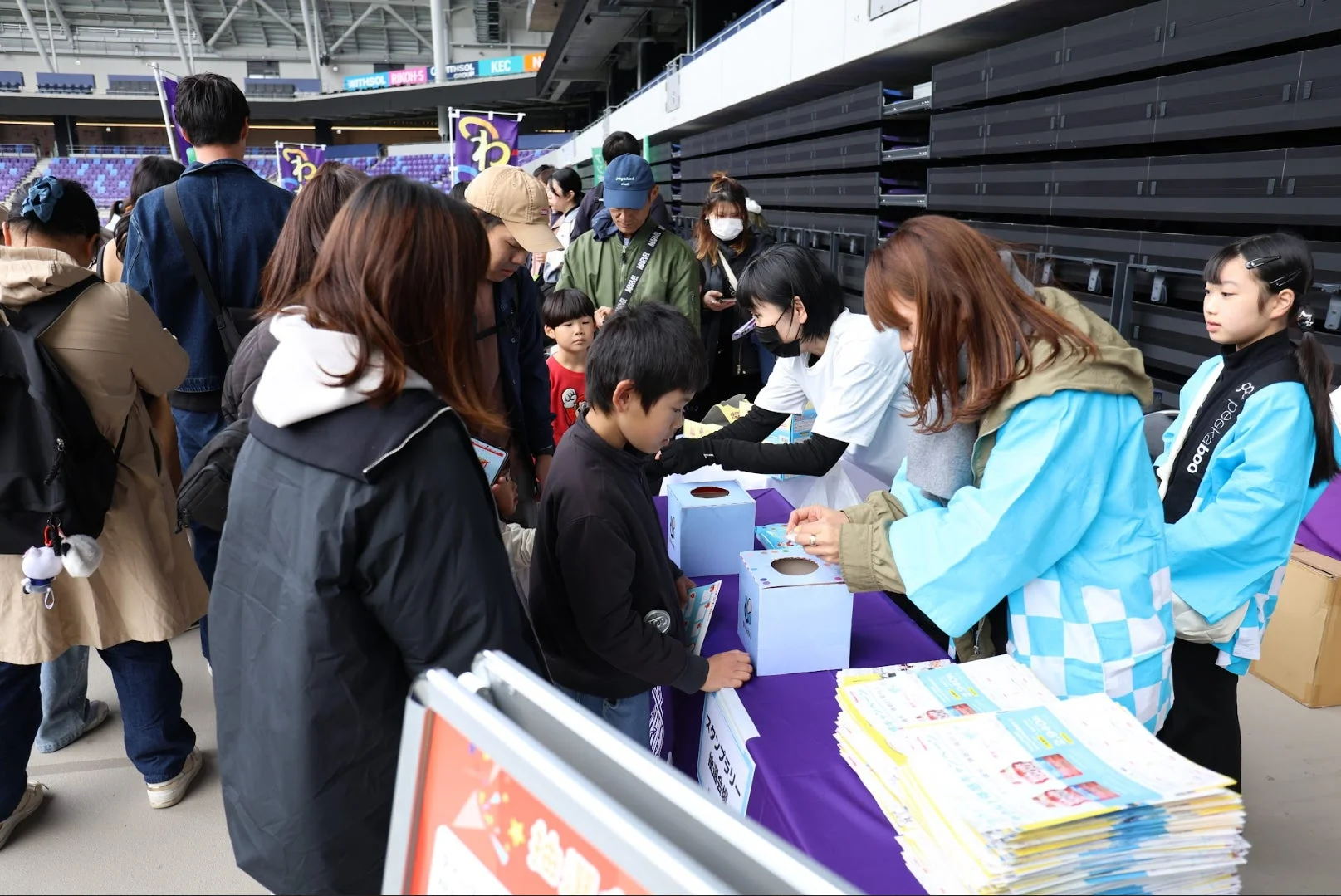 競技場のような広い会場で、多くの人々がイベントに参加している様子です。スタッフが子供たちに何かを配布しており、スタンプラリーが行われているようです。賑やかな交流の場面が捉えられています。