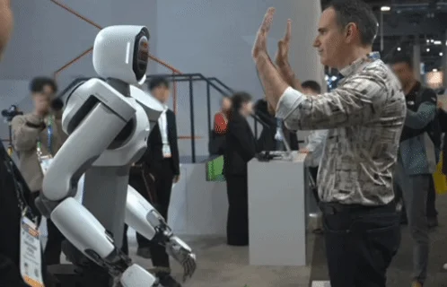 展示会場で、白いヒューマノイドロボットと男性が向かい合い、男性が両手を上げている場面です。人間とロボットの交流や、未来のテクノロジーを示すイベントの一コマと見られます。