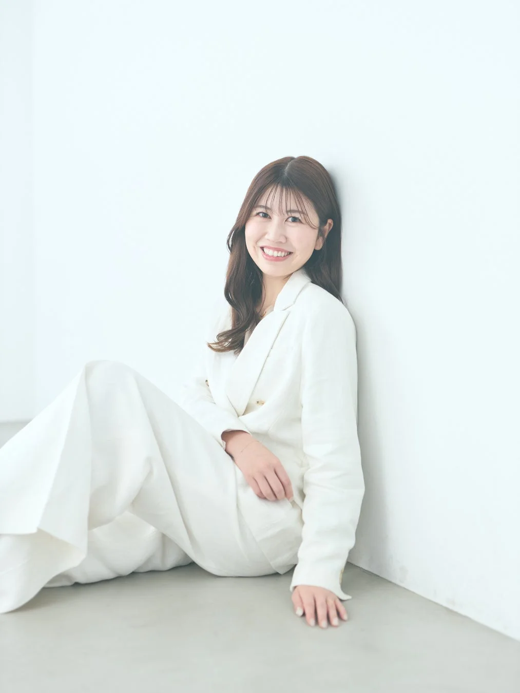 白いスーツを着た若い女性が、白い壁にもたれて床に座り、カメラに向かって明るく微笑んでいるポートレート写真