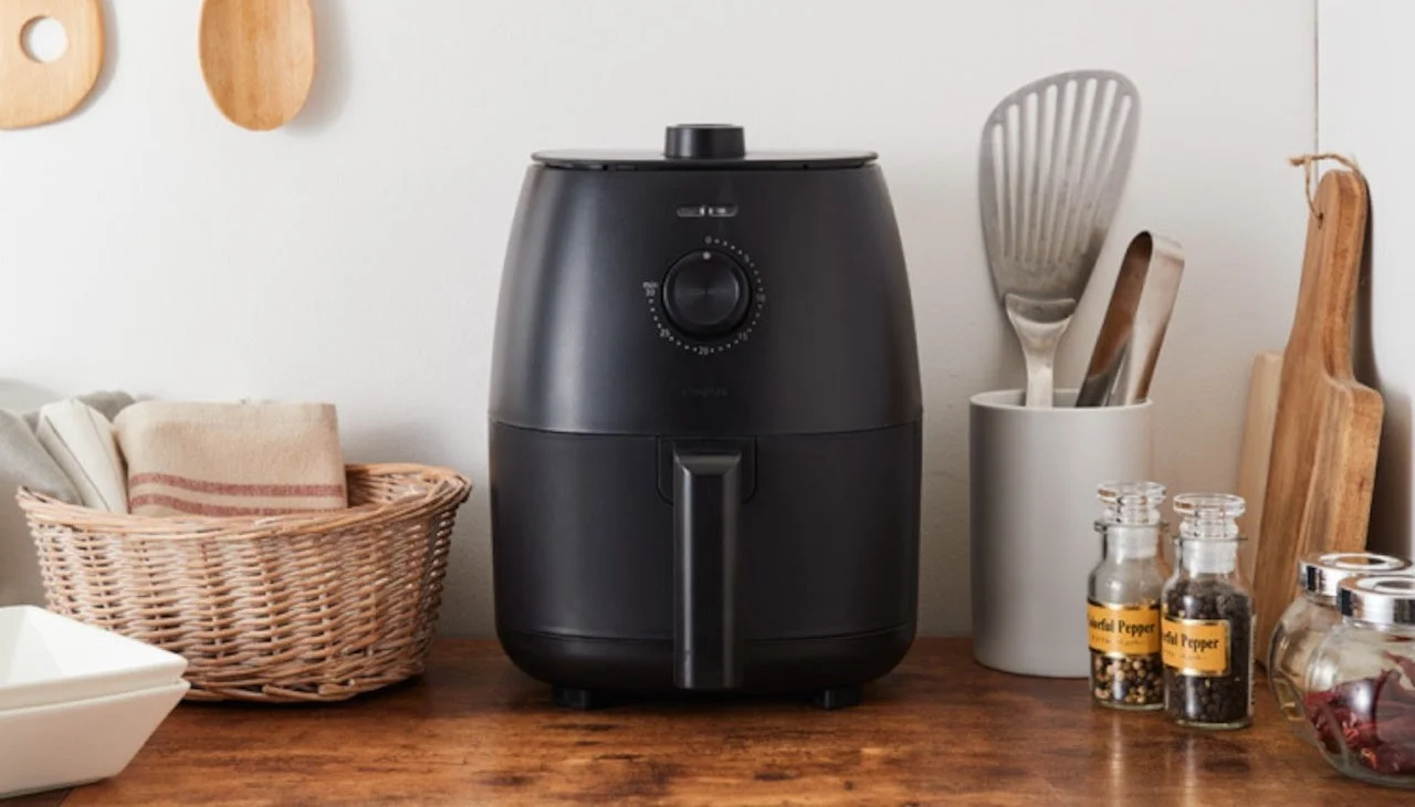 木のカウンターに置かれた黒いエアフライヤーが中心のキッチン風景。周りにはカゴに入った布や調理器具、スパイス瓶が並べられ、すっきりと整頓された印象を与える。