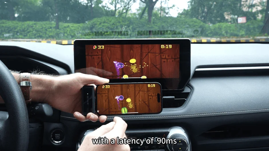 車内でスマートフォンをインフォテインメントシステムにミラーリングし、ゲームをプレイしている様子。画面には「遅延90ms」と表示されており、スマートフォンと車載ディスプレイの表示性能を比較している状況を示しています。