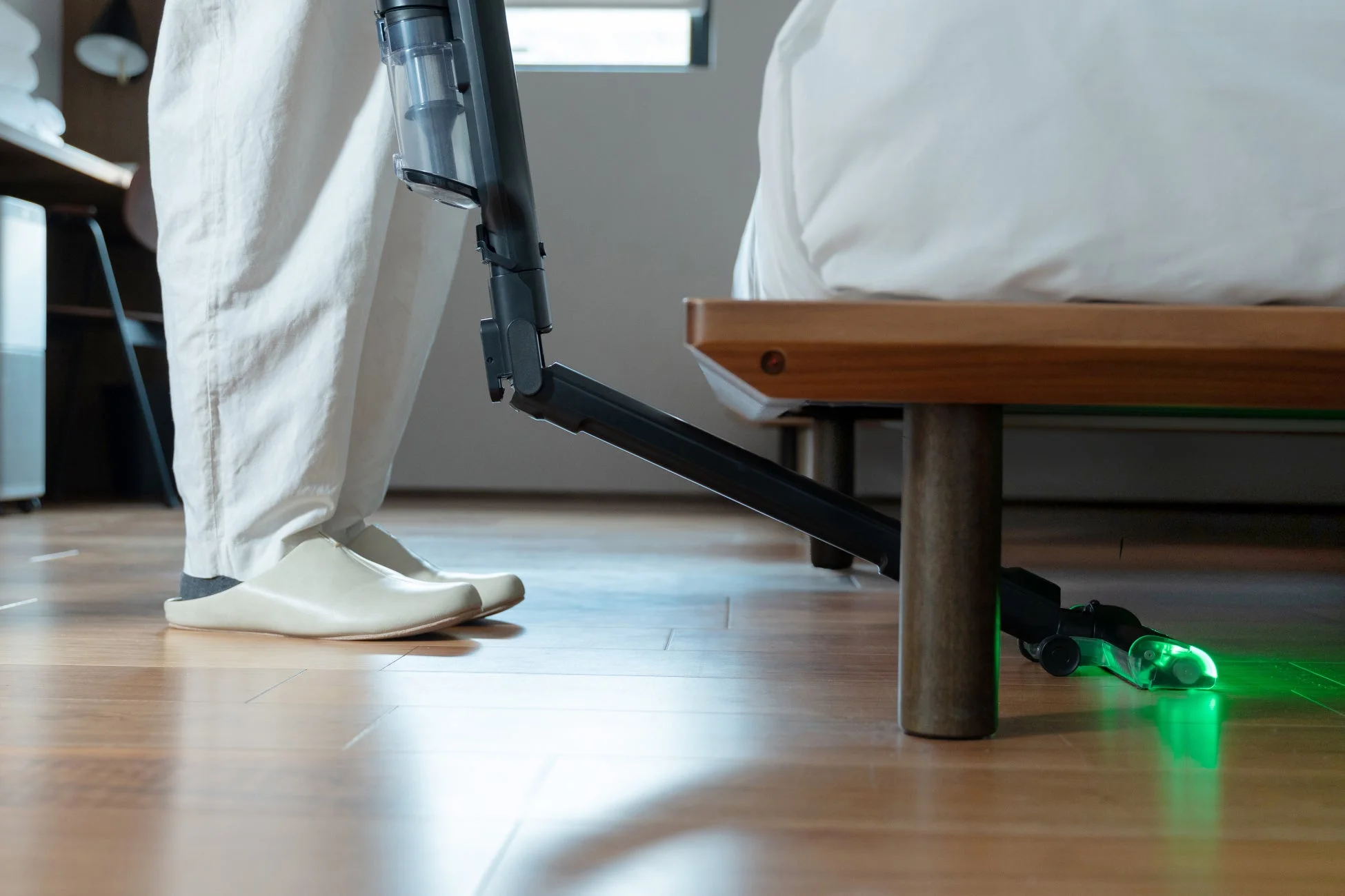 ベッドの下を掃除する様子。スティッククリーナーのヘッドから緑色の光が照射され、暗い場所のゴミを照らしている