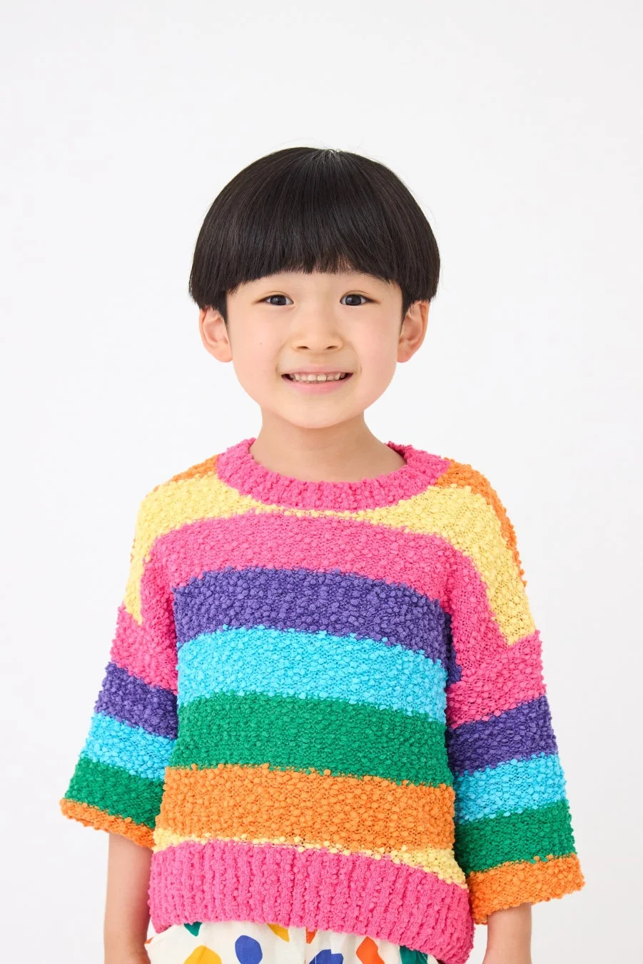 白い背景の前で、カラフルなレインボー柄のセーターを着たアジア人の子供が笑顔でカメラを見つめているポートレート写真です。明るく元気な印象を与えます。