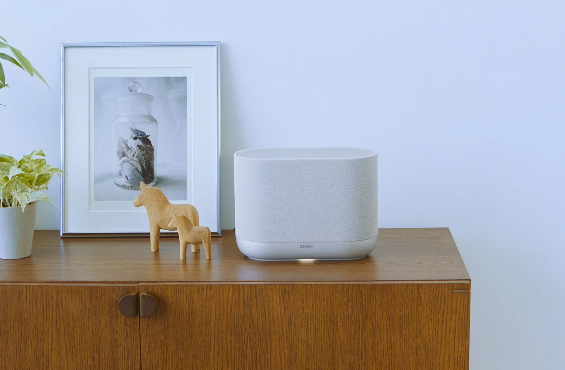 木製のキャビネットの上に、デノンのワイヤレススピーカー、観葉植物、木製の馬の置物、そして鳥の絵が飾られた額縁が配置されています。すっきりと整えられたモダンなインテリア空間です。