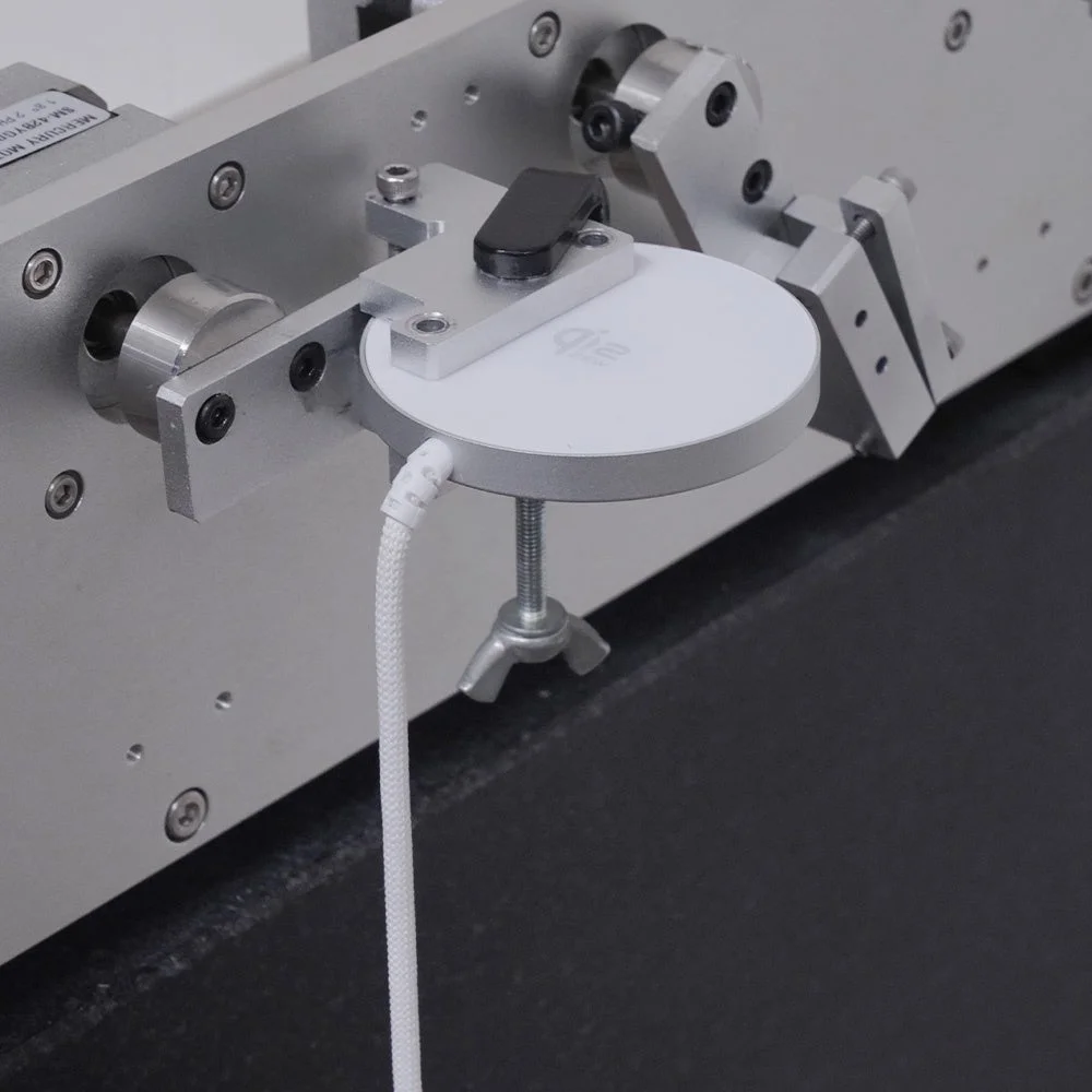 白い円形のデバイスが金属製の機械装置に固定され、白い編組ケーブルで接続されている様子です。何らかの試験や測定が行われている精密機器の一部を示しています。