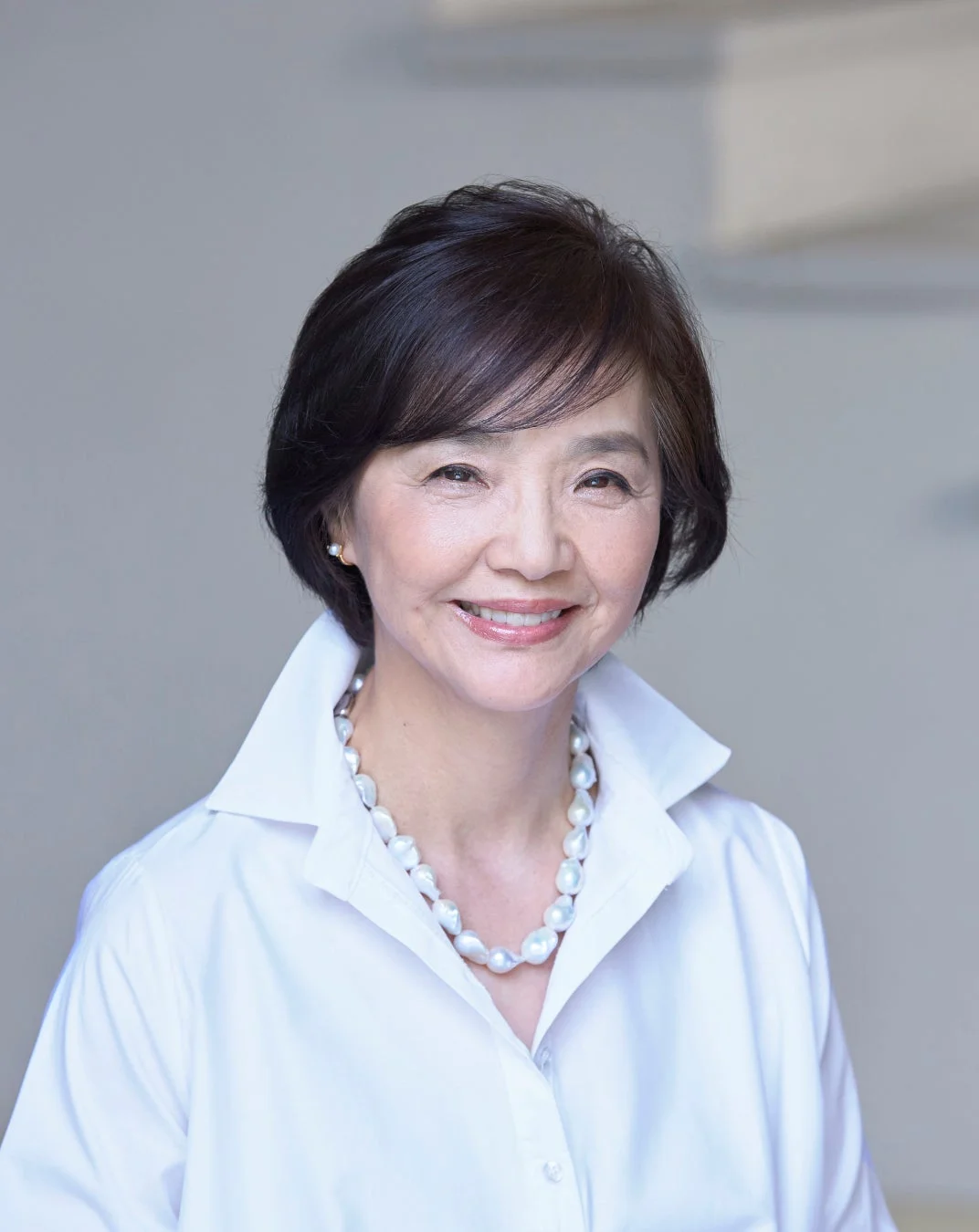 白いシャツとパールのネックレスを身につけ、カメラに向かって優しく微笑むショートヘアの女性のポートレートです。