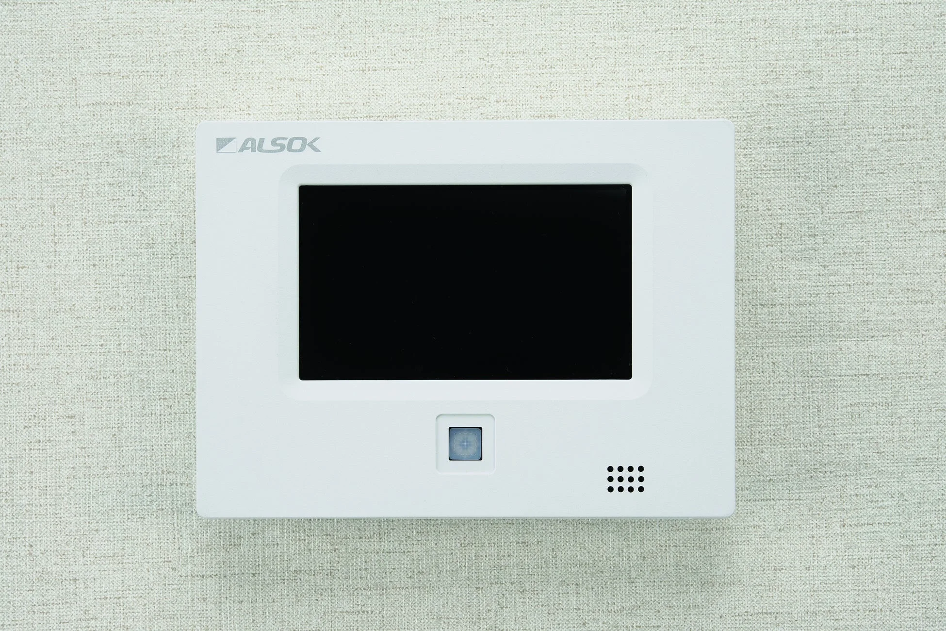 白い壁に取り付けられたALSOKのセキュリティパネルまたはインターホンです。黒い画面、中央のボタン、右下のスピーカーグリルが特徴で、室内の防犯システムの一部として機能する機器と考えられます。