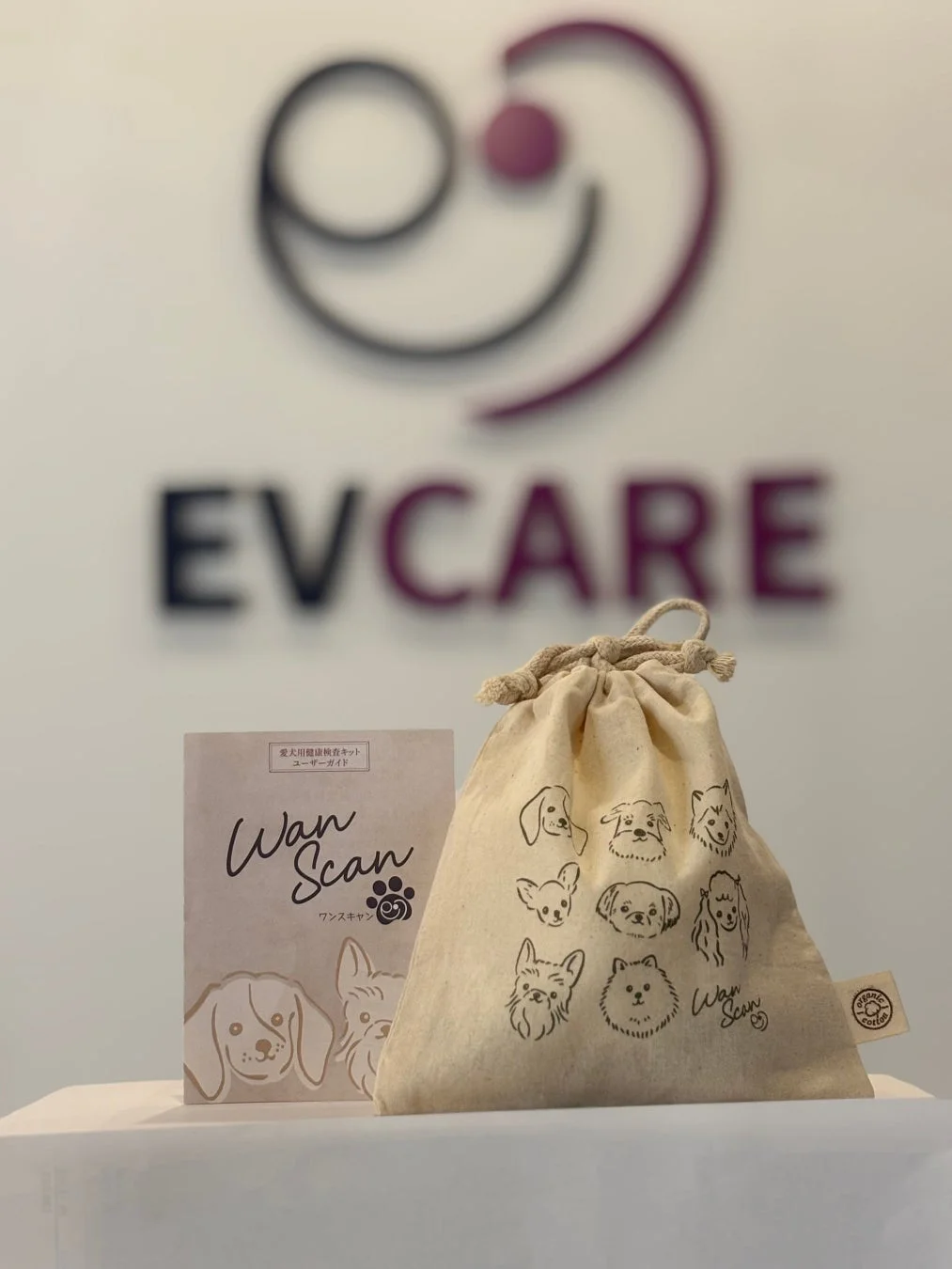 「EVCARE」のロゴがぼやけた背景に、愛犬健康検査キット「Wan Scan」のユーザーガイドと、犬のイラストが描かれたオーガニックコットン製の巾着袋が置かれている画像です。犬の健康管理製品を示すセット内容が写っています。