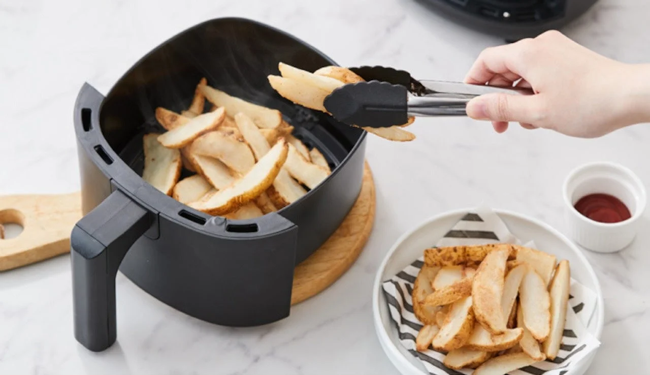 エアフライヤーで調理されたポテトウェッジを、トングを使ってお皿に移している様子です。白いお皿にはストライプ柄の紙が敷かれ、ケチャップの小皿も添えられています。ヘルシーな調理風景を捉えた一枚です。