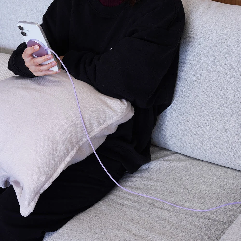 ソファに座り、スマートフォンを操作している人物の様子を捉えた画像です。白いスマートフォンには紫色のMagSafeアクセサリーが装着されており、そこから伸びる紫色のケーブルが特徴的です。自宅でのリラックスしたスマートフォンの利用シーンが描かれています。