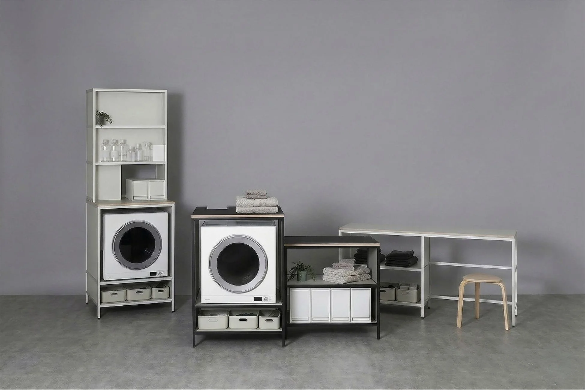 グレーの壁と床の空間に、洗濯機と乾燥機を中心としたモダンで機能的なランドリースペースが配置されています。白い棚や黒いフレームの収納ラックにタオルや洗剤ボトルが整理され、シンプルな木製スツールも置かれています。