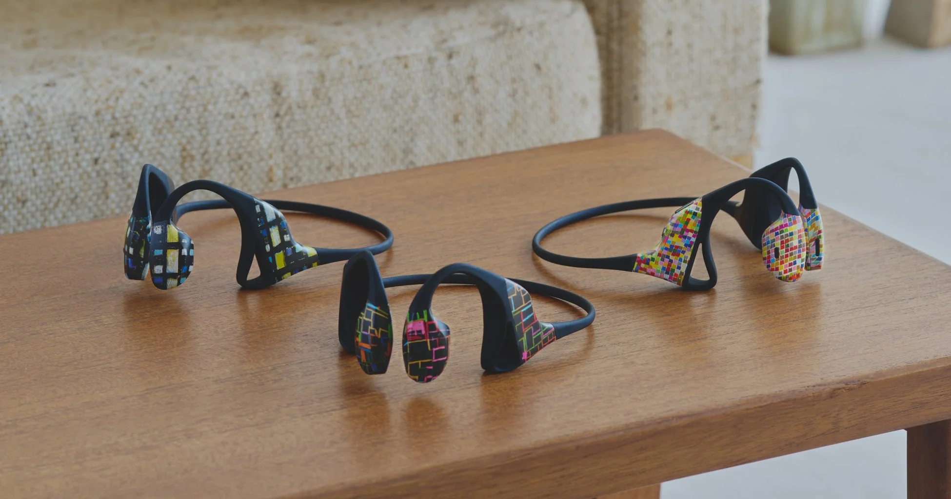 木製のテーブルに、カラフルな柄が施された3つの骨伝導ワイヤレスイヤホンが並べられています。