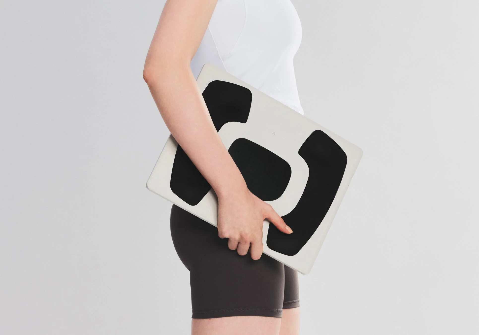 白い背景の前で、女性が体組成計を抱えている横向きのショットです。健康や体重管理のイメージを伝えます。