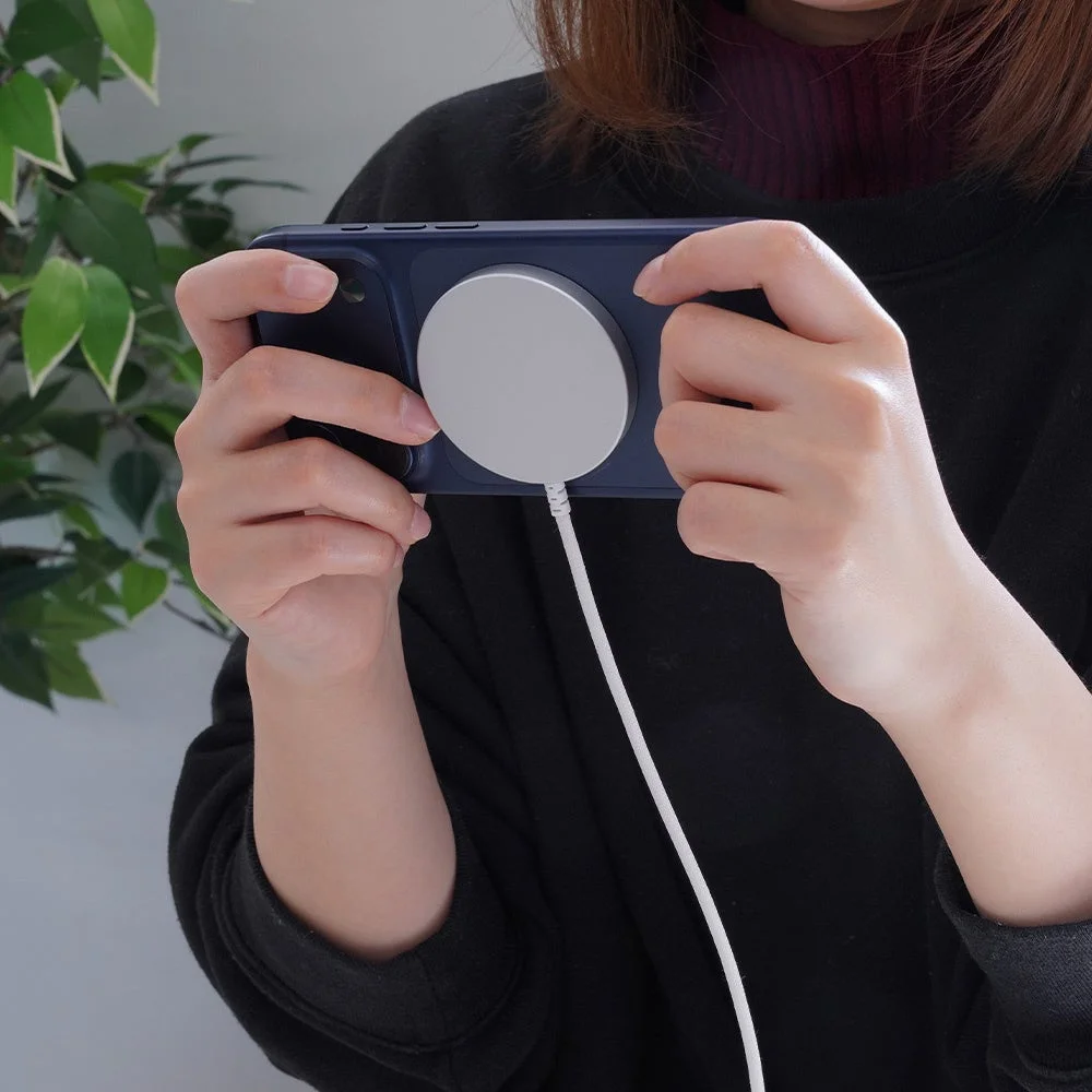 スマートフォンを横向きに持ち、背面に磁気充電器を装着している人物の手元が写っています。充電中のスマートフォン操作を表すシーンです。
