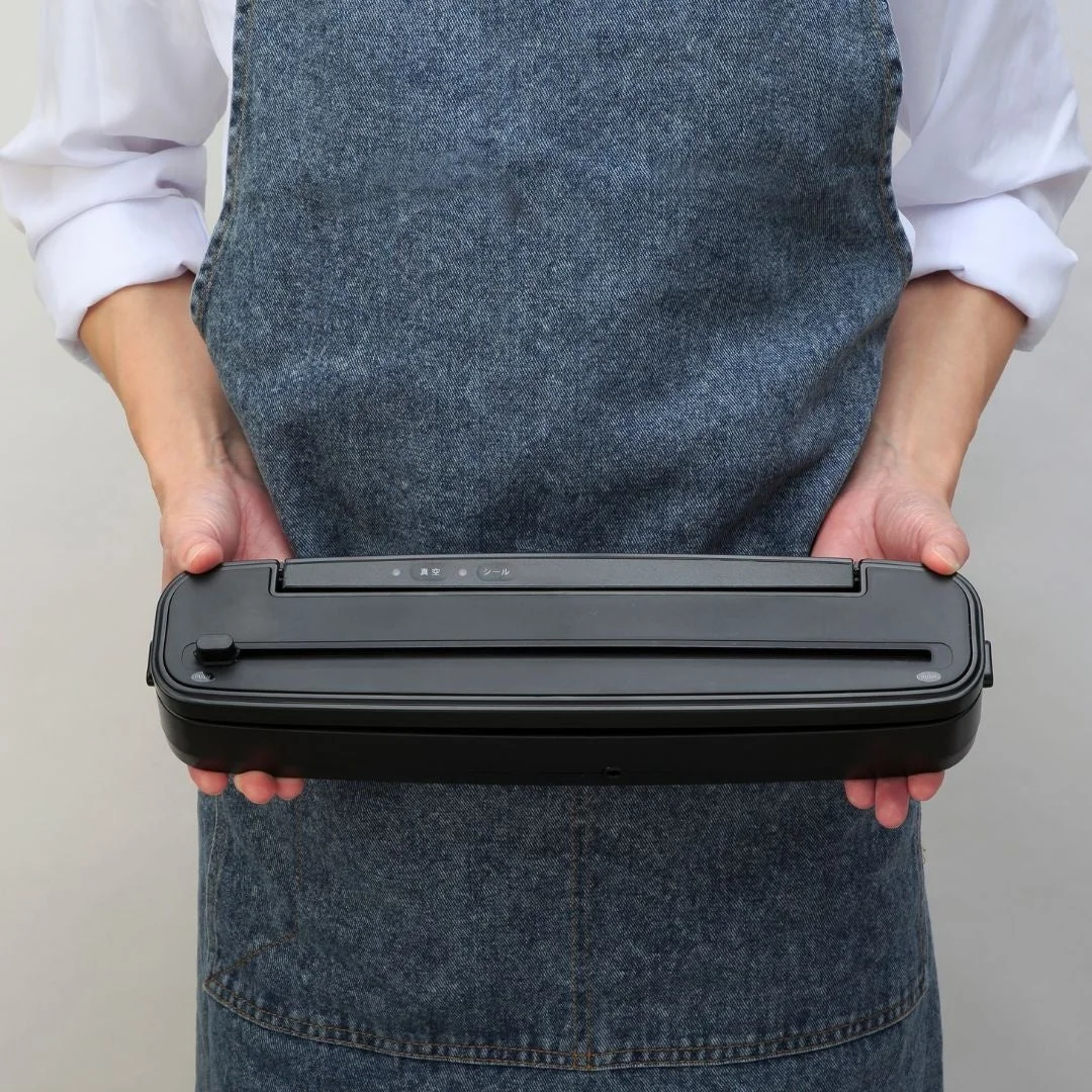 デニムエプロンを着用した人物が、両手で黒い真空パック器を水平に持っている画像です。
