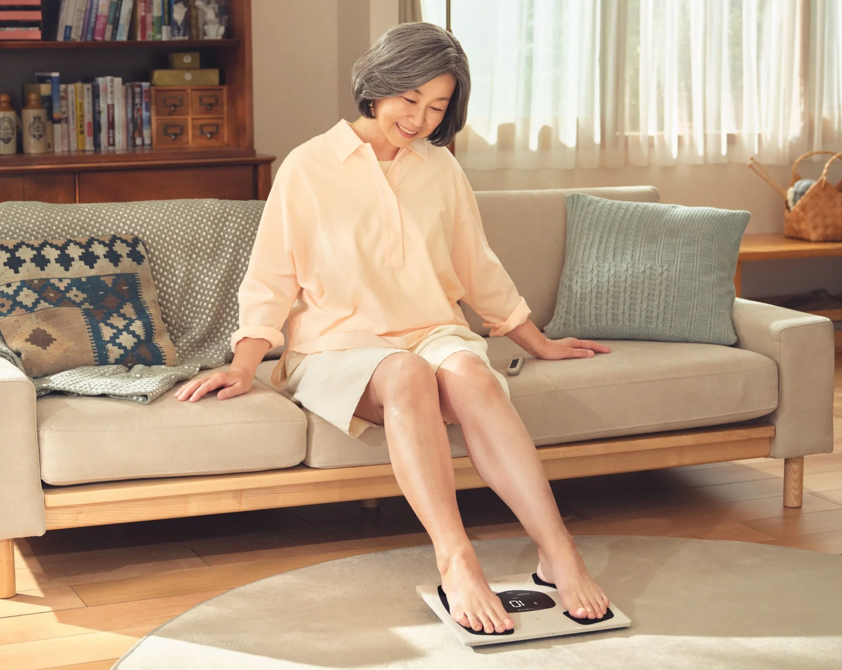 リビングのソファに座った笑顔のシニア女性が、スマート体重計で健康状態を測定しています。健康的なライフスタイルを過ごす日常の一コマが描かれています。
