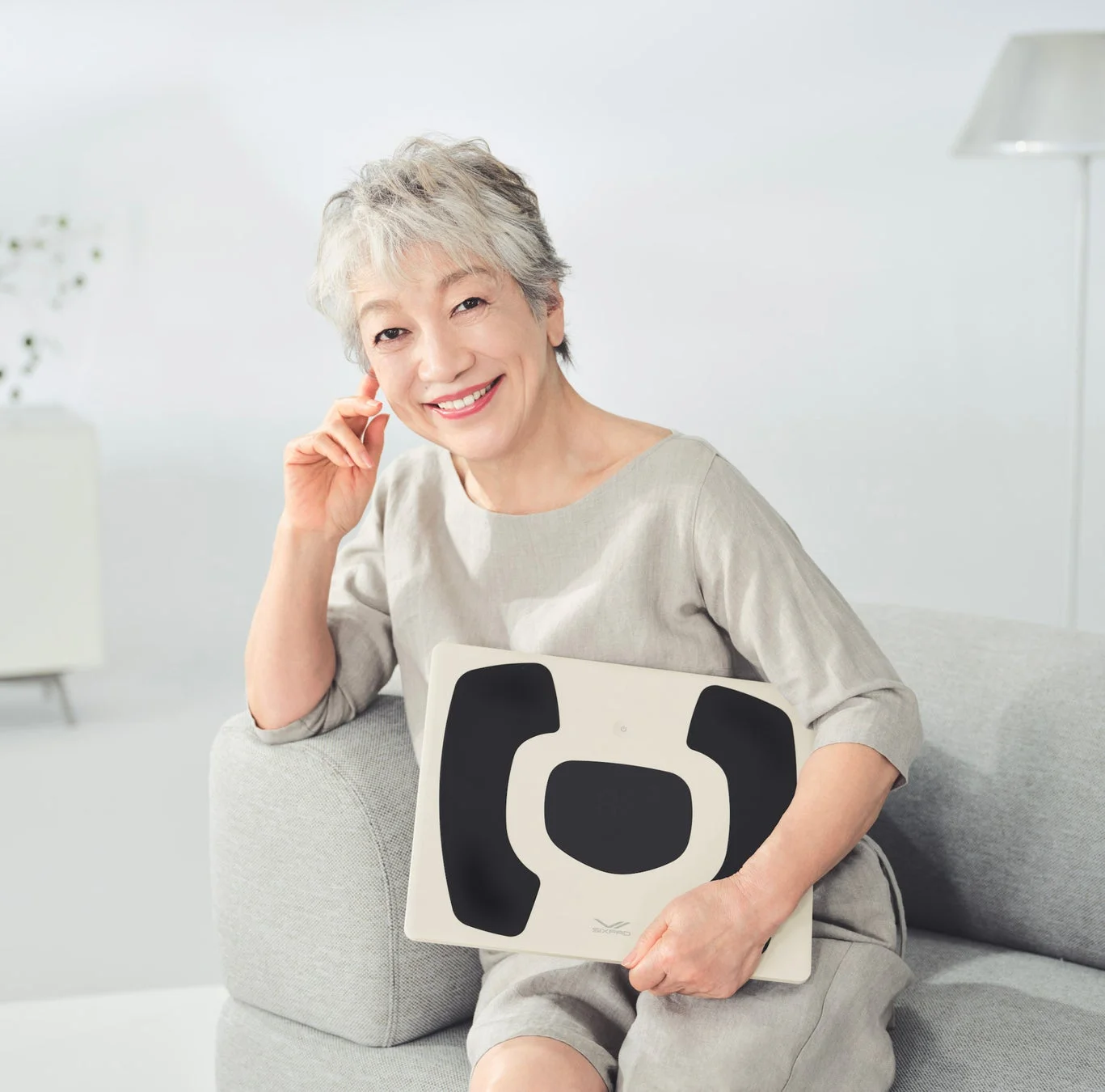 グレーのソファに座り、笑顔でカメラを見つめる高齢の女性。彼女はSIXPADの体組成計を手に持ち、リラックスした様子です。明るくモダンなリビングルームが背景にあり、健康的なライフスタイルを想起させます。