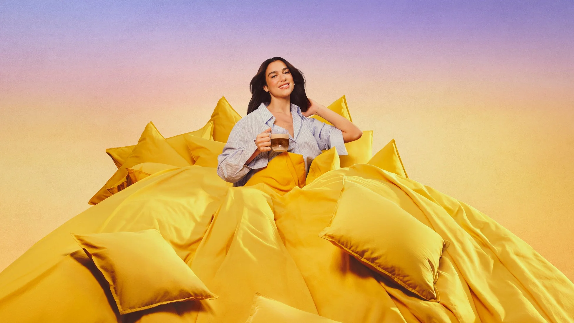 ストライプシャツを着たデュア・リパがベッドでコーヒーを手に笑顔を見せる様子