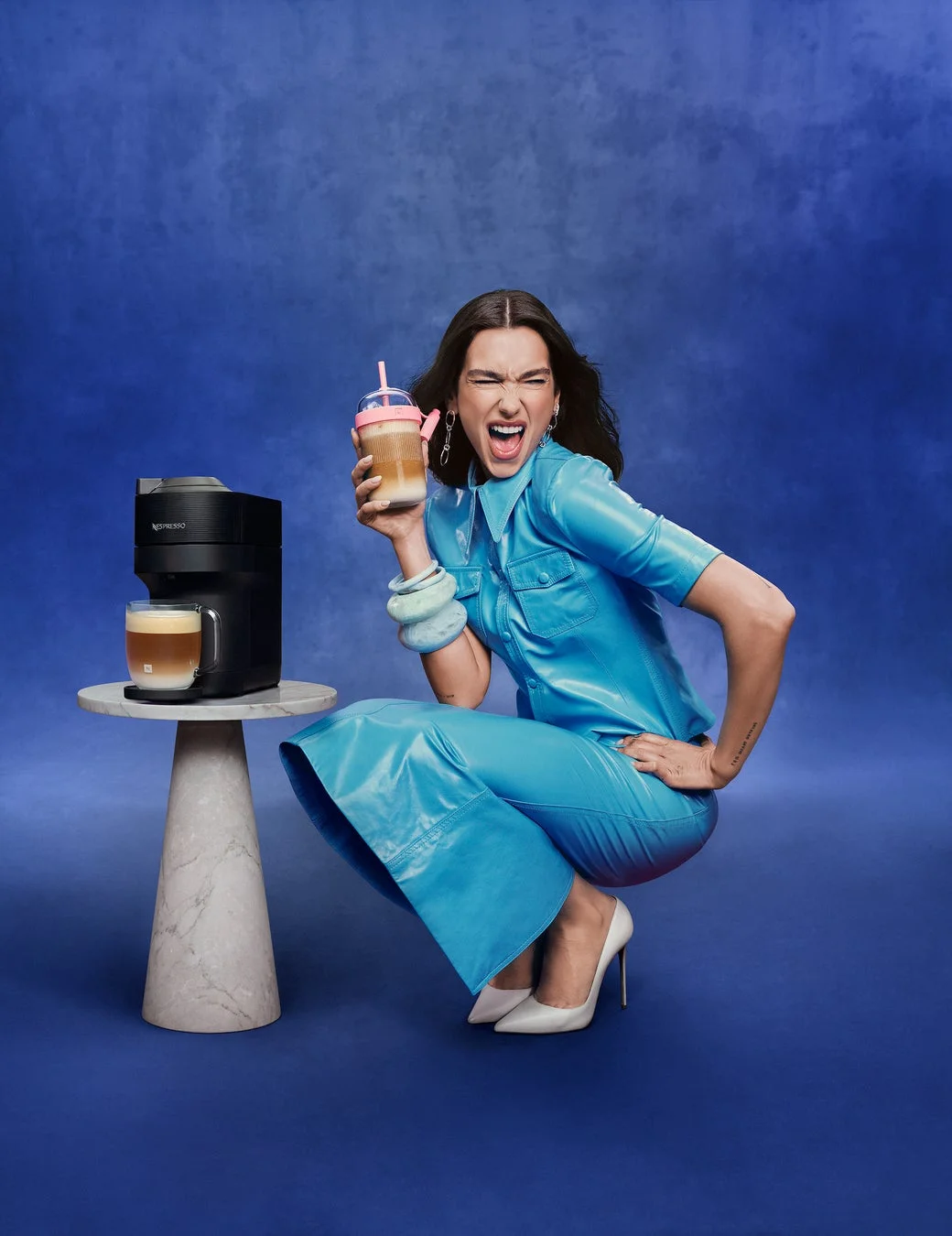 青いレザースーツを着たデュア・リパがネスプレッソのコーヒーメーカーの隣でアイスコーヒーを持つ様子