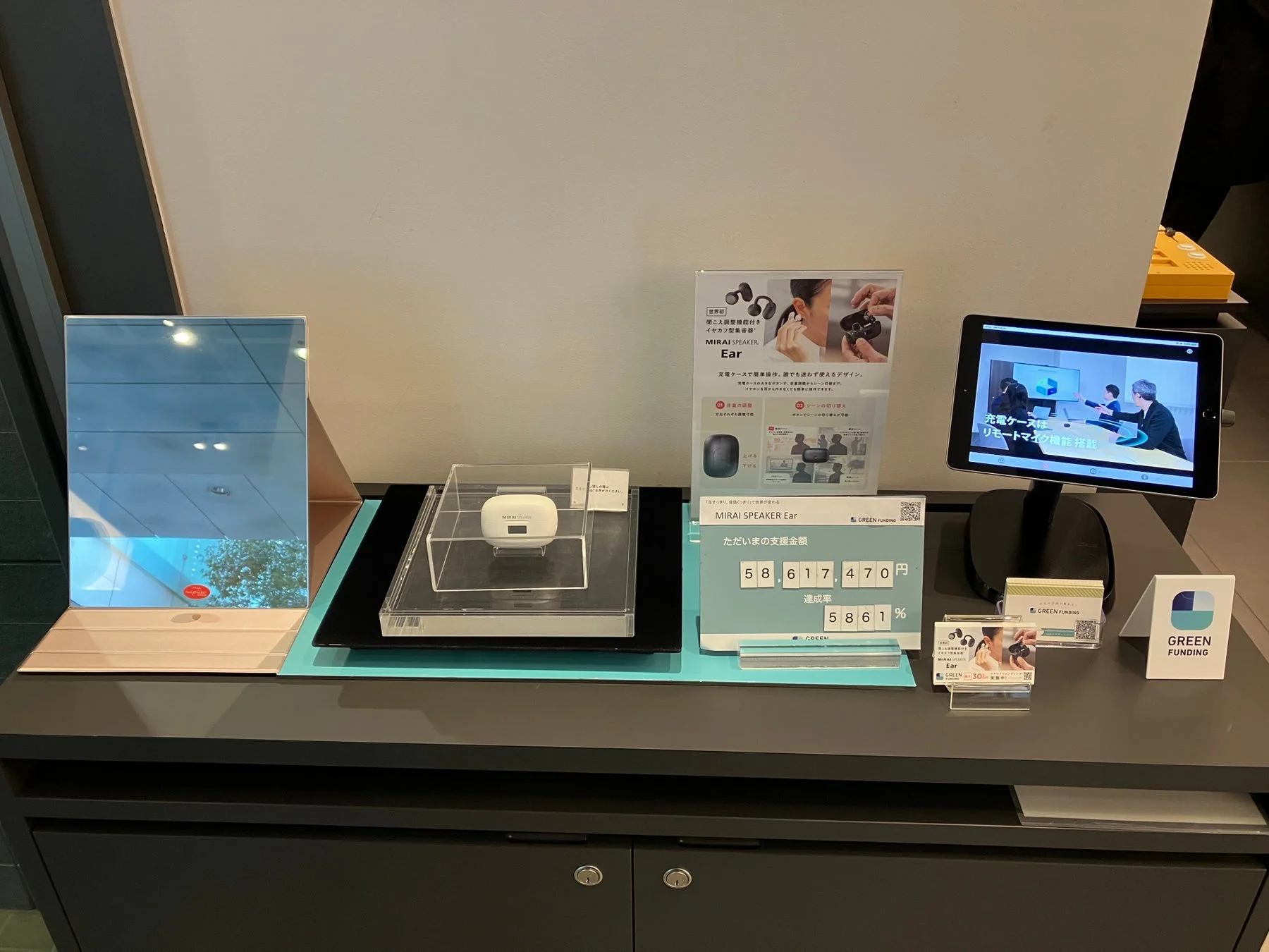 イヤホン型骨伝導スピーカー「MIRAI SPEAKER Ear」の製品展示風景です。クラウドファンディング「GREEN FUNDING」の達成状況（支援金額5,861,470円、達成率5861%）が示され、製品の機能やデザイン、充電ケースのリモートマイク機能について説明されています。