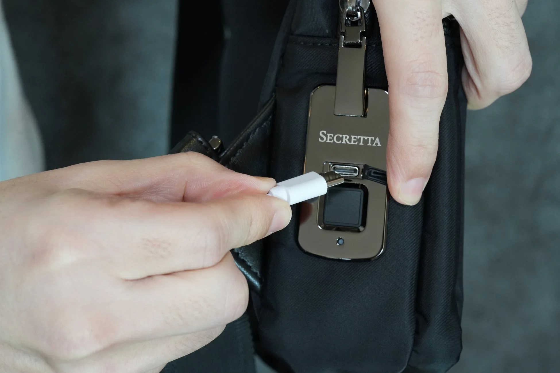 黒いバッグのUSB充電ポートに白いケーブルを差し込んでいる様子。バッグには「SECRETTA」と刻印された金属製のパーツと指紋認証センサーが見える。