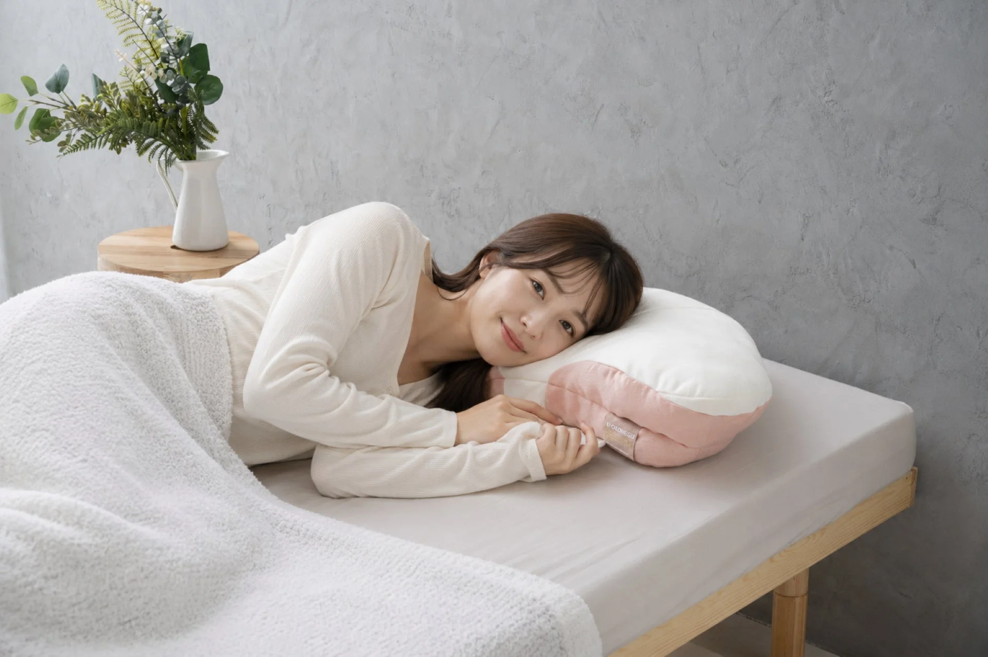 ベッドに横たわり、ピンクと白の枕に頭を乗せて優しく微笑む女性の姿。くつろいだ雰囲気。