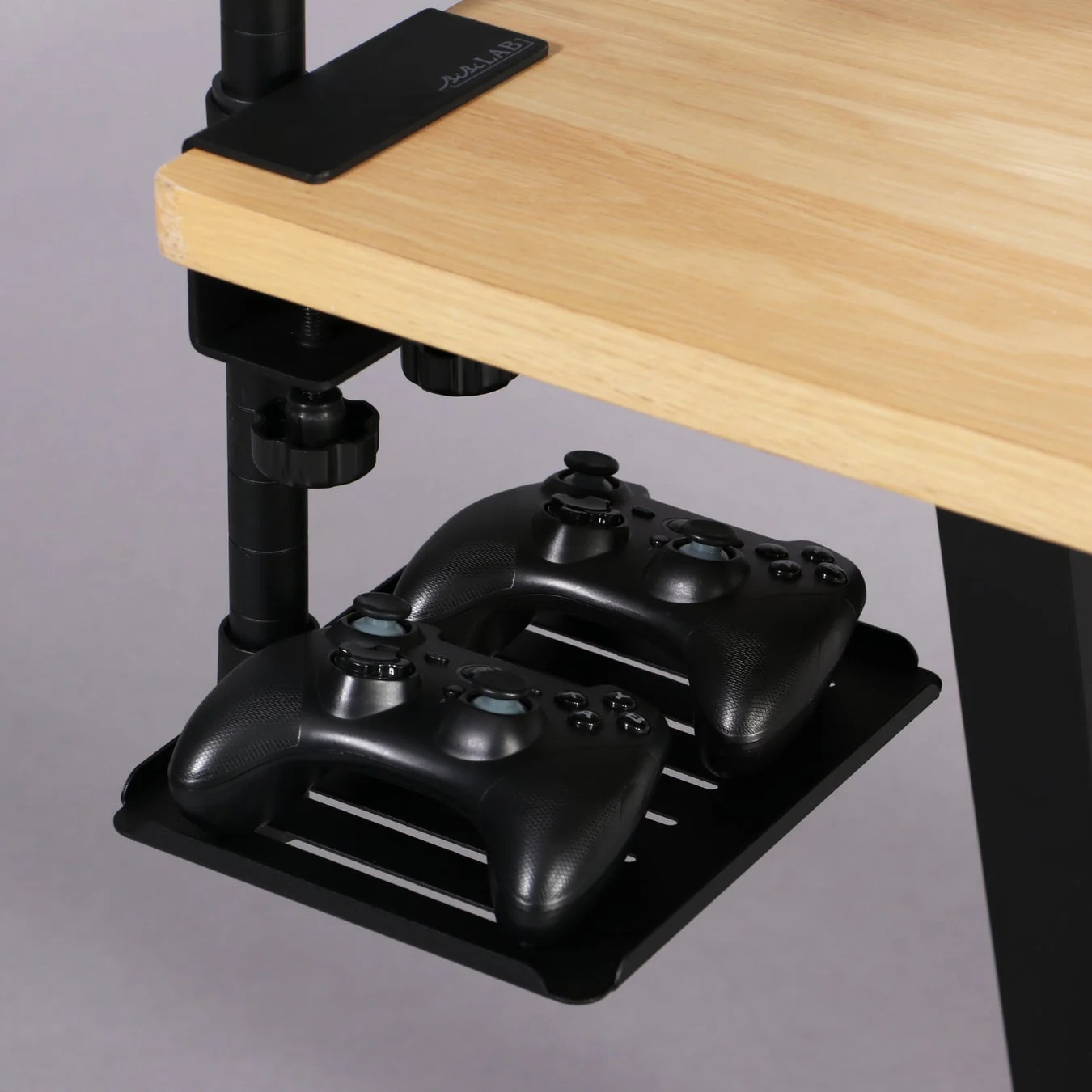 木目のデスクの裏にクランプで取り付けられた黒い棚に、2つのゲームコントローラーが収納されている様子