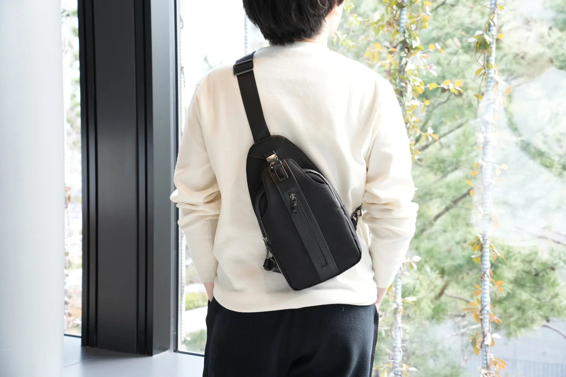 白いセーターと黒いスリングバッグを身につけた男性が、大きな窓から外の緑を眺めている後ろ姿の画像です。