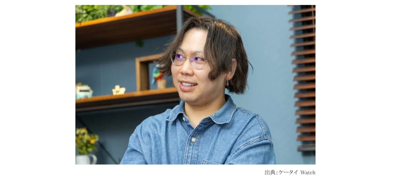 眼鏡をかけたアジア系の男性がデニムシャツを着て、室内で穏やかな笑顔を見せています