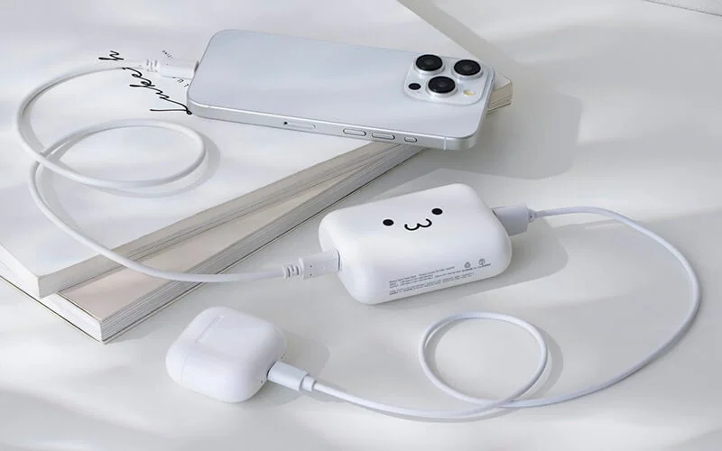 白いスマートフォンとワイヤレスイヤホンケースが、かわいい顔の描かれた白いモバイルバッテリーで同時に充電されています。